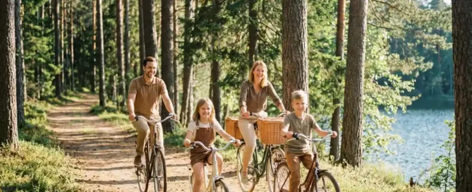 Familie van vier fietst op zonnig bospad bij het Veluwemeer, met rieten picknickmand op de fiets en dennen op de achtergrond.