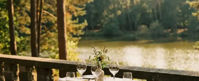 Elegant lunchtafel voor twee op een zonnig terras met uitzicht op een bosmeer in de Veluwe, kristallen glazen en linnen servetten.