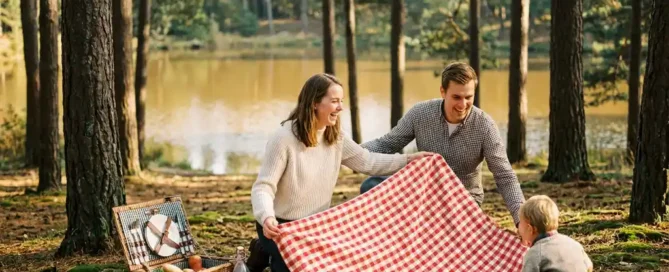 Jong gezin spreidt een geruite picknickdeken op de bosgrond van de Veluwe, met een rieten mand vol sandwiches en fruit bij een meer.