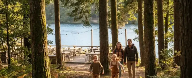 Familie met jonge kinderen op een bospad op de Veluwe, houten restaurantterras en glinsterende meer zichtbaar tussen de dennenbomen.