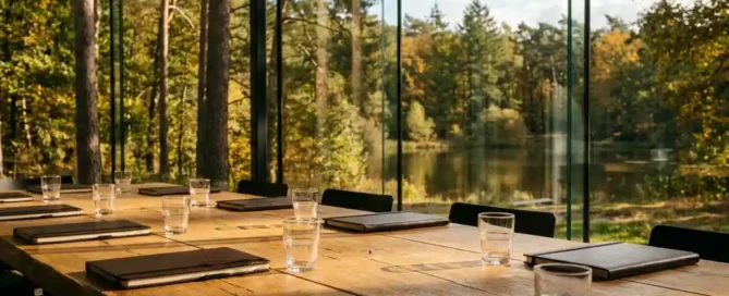 Eikenhouten vergadertafel met leren notitieboeken bij panoramaramen met uitzicht op het Veluwse bos en meer in herfstig goudlicht.